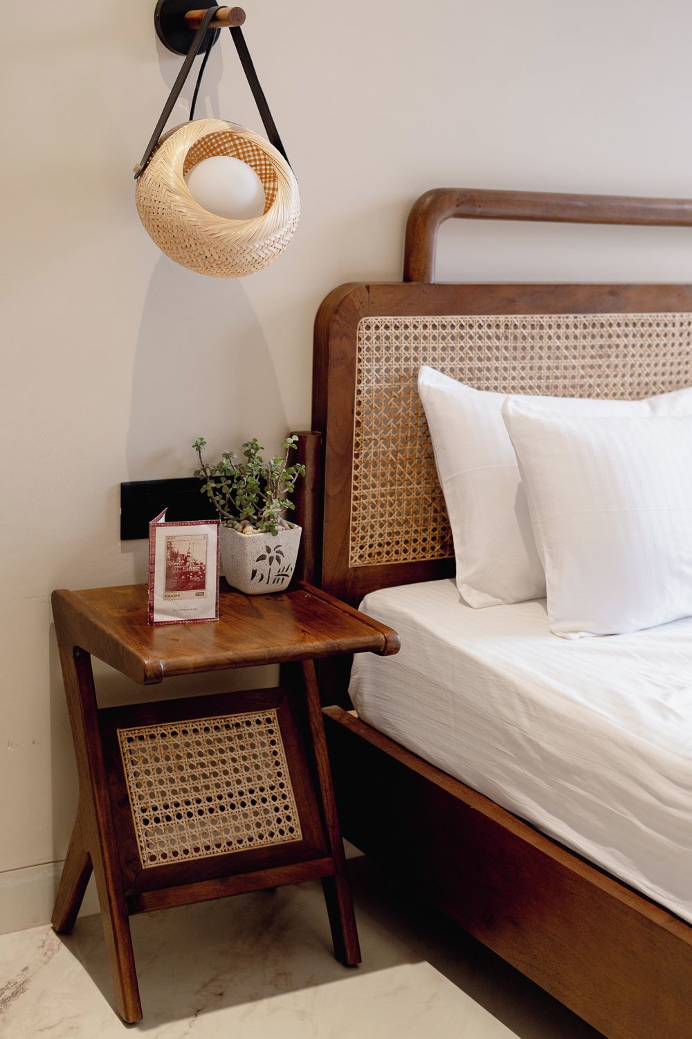 A wooden bed with a woven headboard, a small wooden nightstand with a woven basket, a potted plant, and a framed picture.