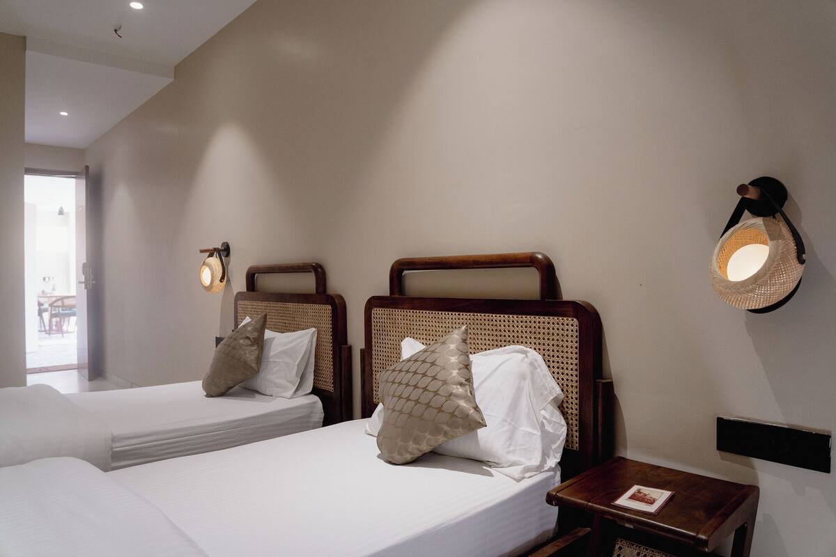 A hotel room with two single beds, a small bedside table with a book, a wall-mounted lamp, and a view of the outside.