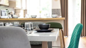 Un comedor con una mesa lista para dos, una silla tapizada de verde y una cocina al fondo.
