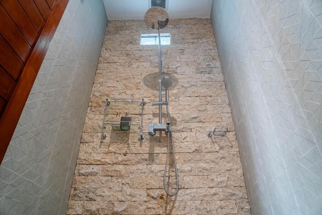A bathroom with a stone floor, a shower with a handheld showerhead, and a wall-mounted fan.