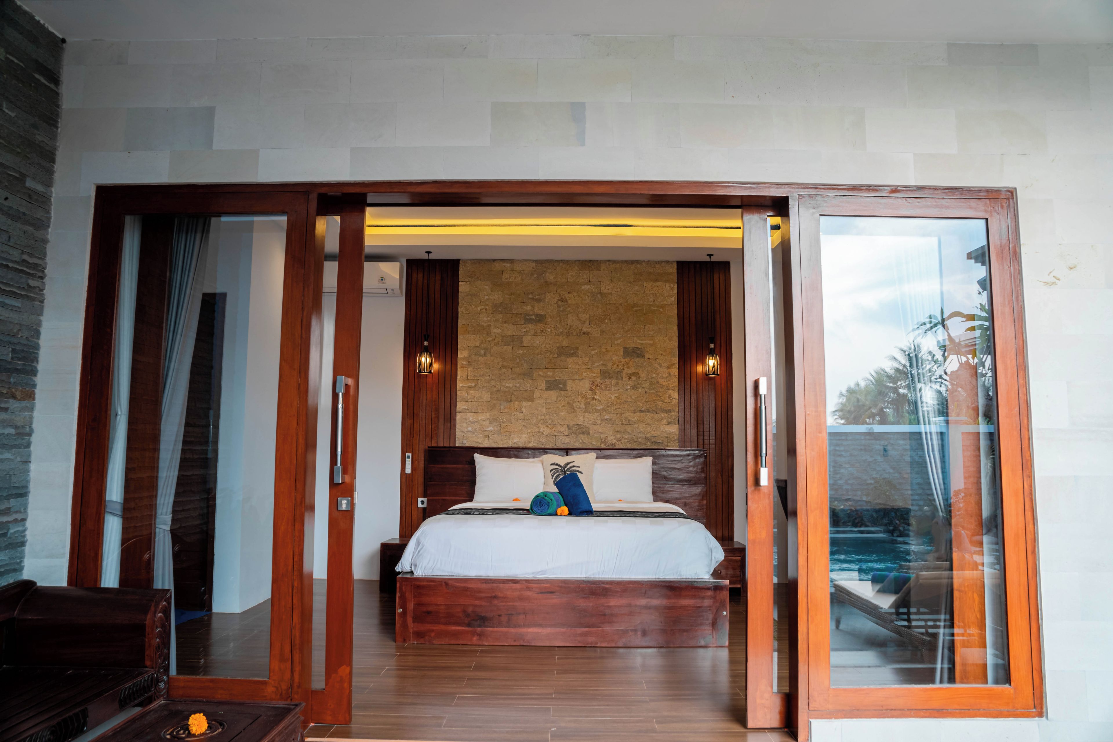 A bedroom with a wooden bed, a stone wall, and a view of a pool through a glass door.
