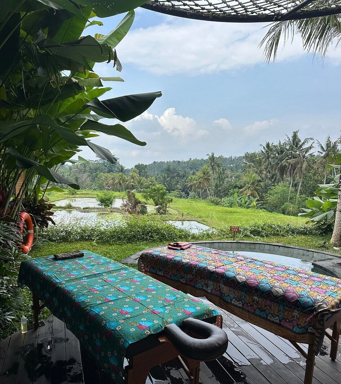 Two massage tables with colorful covers, set on a wooden deck with a view of a lush landscape and rice paddies.