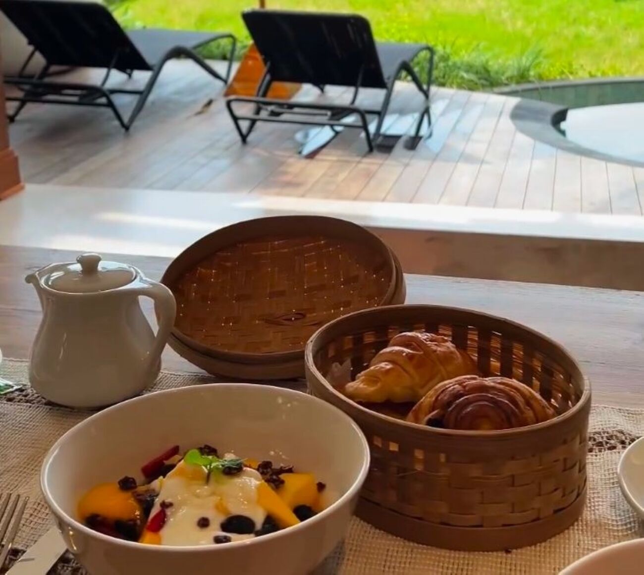Una colazione con una ciotola di frutta, un cestino di croissant e un teiera su un tavolo con vista su posti a sedere all'aperto.