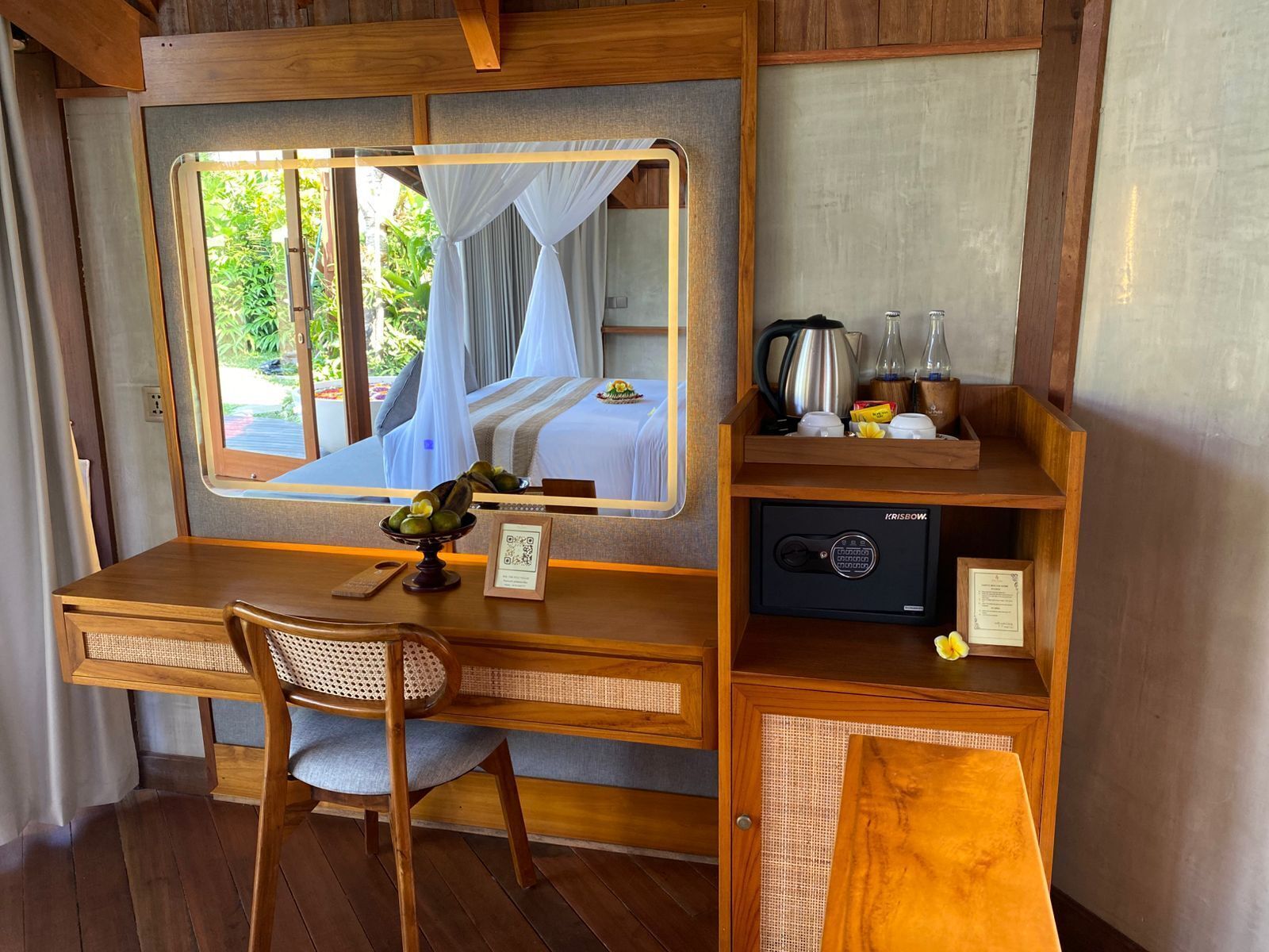 A compact room with a wooden desk, a chair, a bed with a canopy, a window with curtains, a small shelf with kitchen items, and a QR code on the desk.