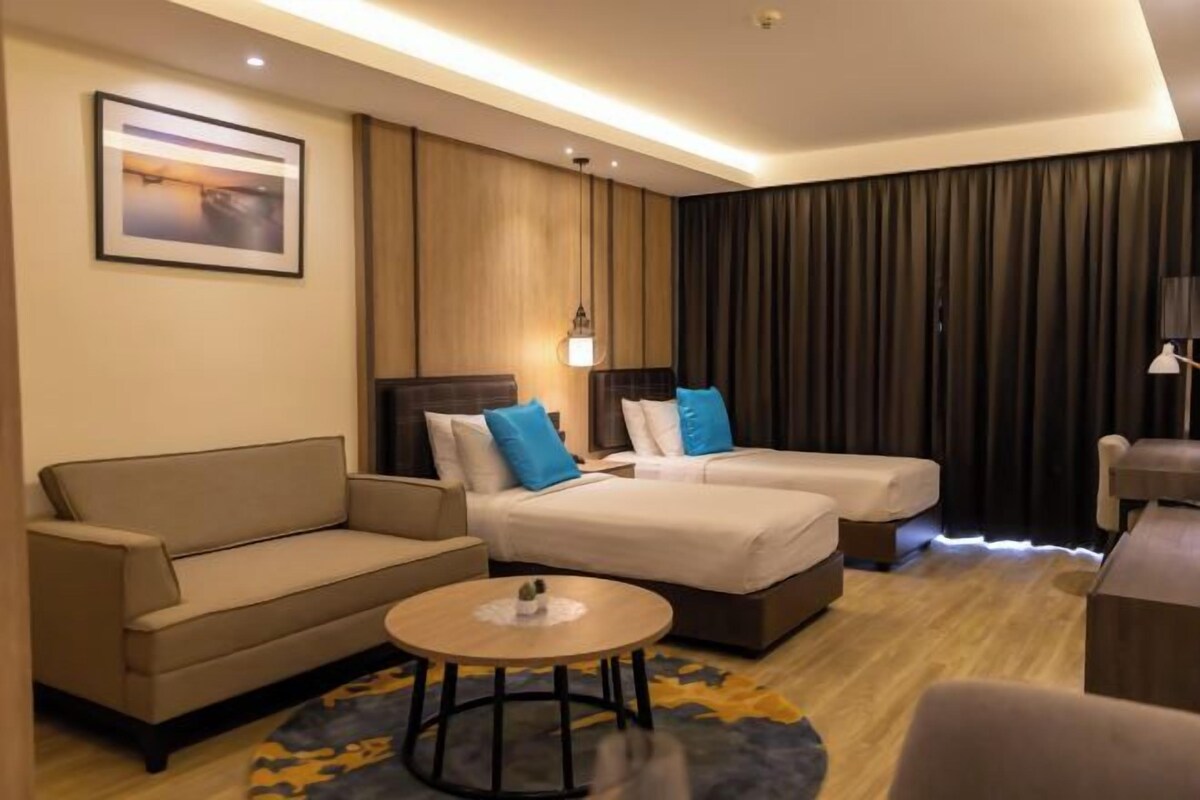 A hotel room with a sofa, two beds with pillows, a coffee table, a lamp, and a framed picture on the wall.