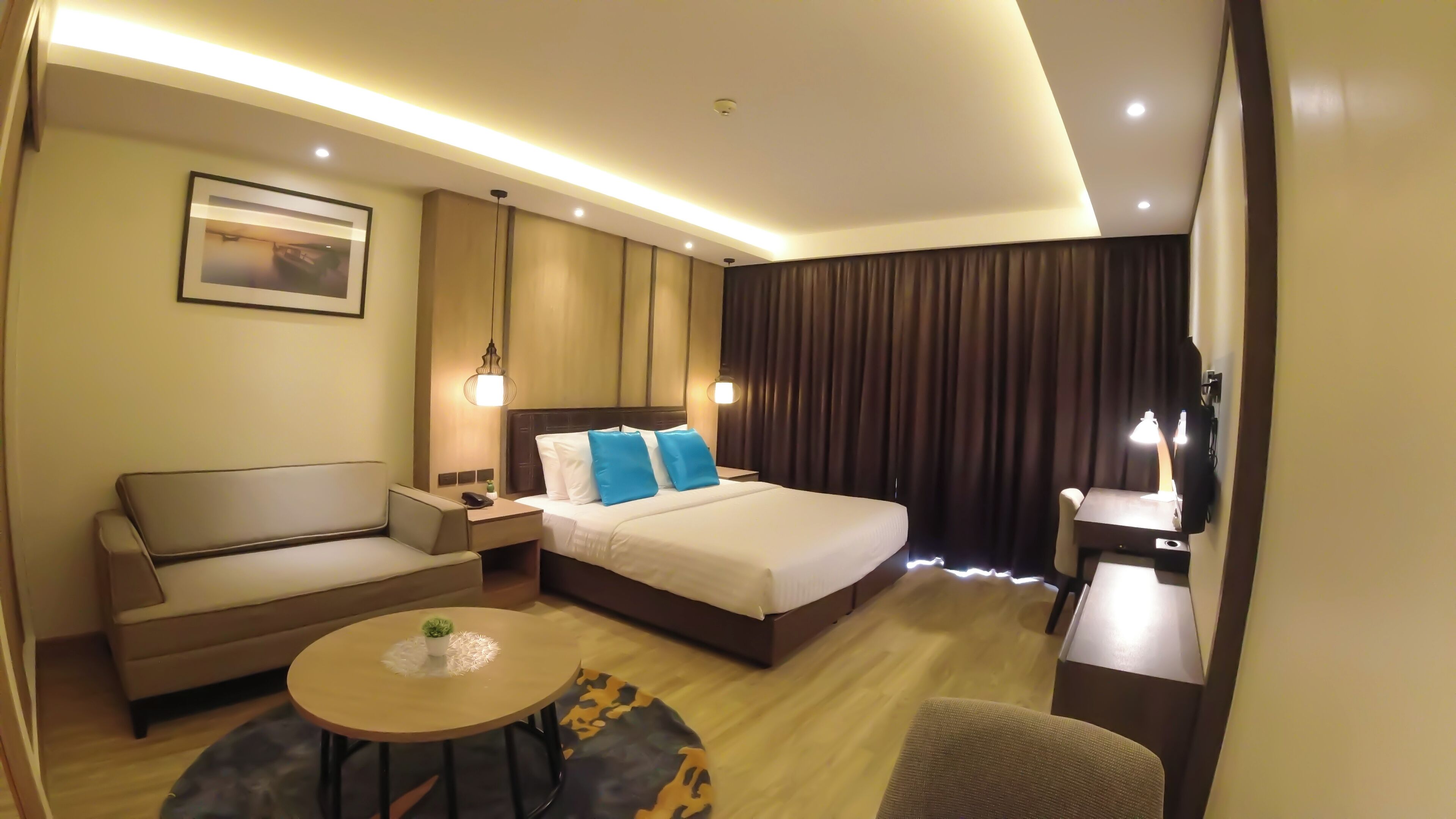A hotel room with a bed, a sofa, a small table with a plant, a desk with a lamp, and a TV.