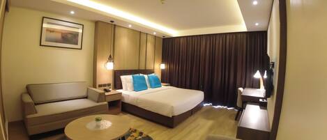 A hotel room with a bed, a sofa, a small table with a plant, a desk with a lamp, and a TV.