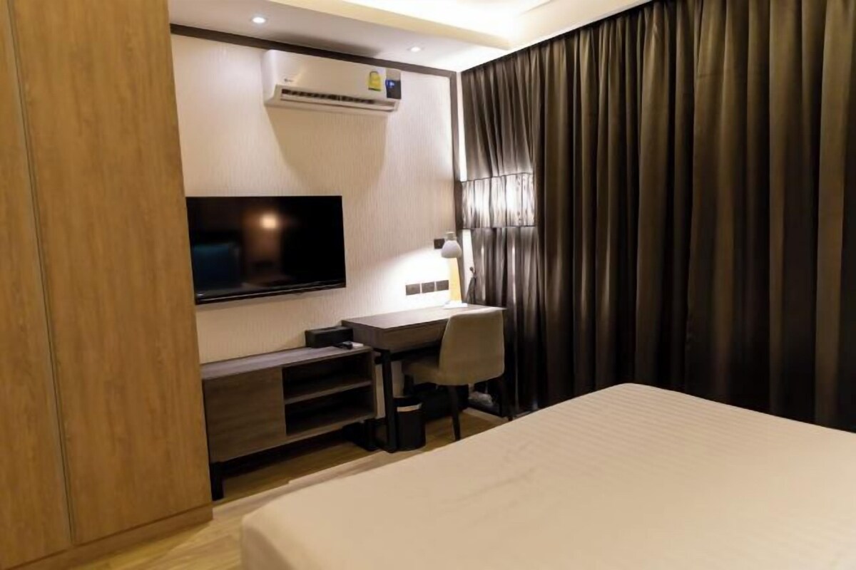 A hotel room with a bed, a desk with a chair, a flat-screen TV, an air conditioning unit, and a large window with curtains.