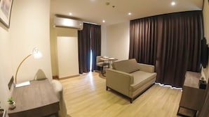 A modern hotel room with a sofa, a small table, a desk with a lamp, an air conditioning unit, and a large window with curtains. - Wyatt Hotel Jomtien Beach (Pattaya)