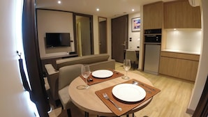 A modern dining area with a round table set for two, a television mounted on the wall, and a kitchen area with wooden cabinets and a refrigerator. - Wyatt Hotel Jomtien Beach (Pattaya)