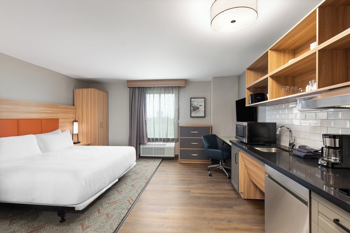 A modern hotel room with a bed, a desk, a kitchenette, and a bathroom.