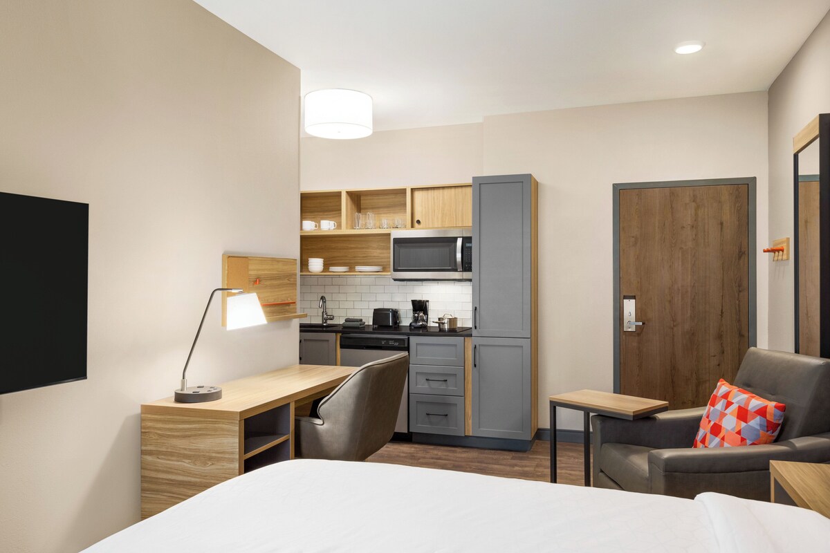 A compact hotel room with a kitchenette, a bed, a desk, and a seating area.