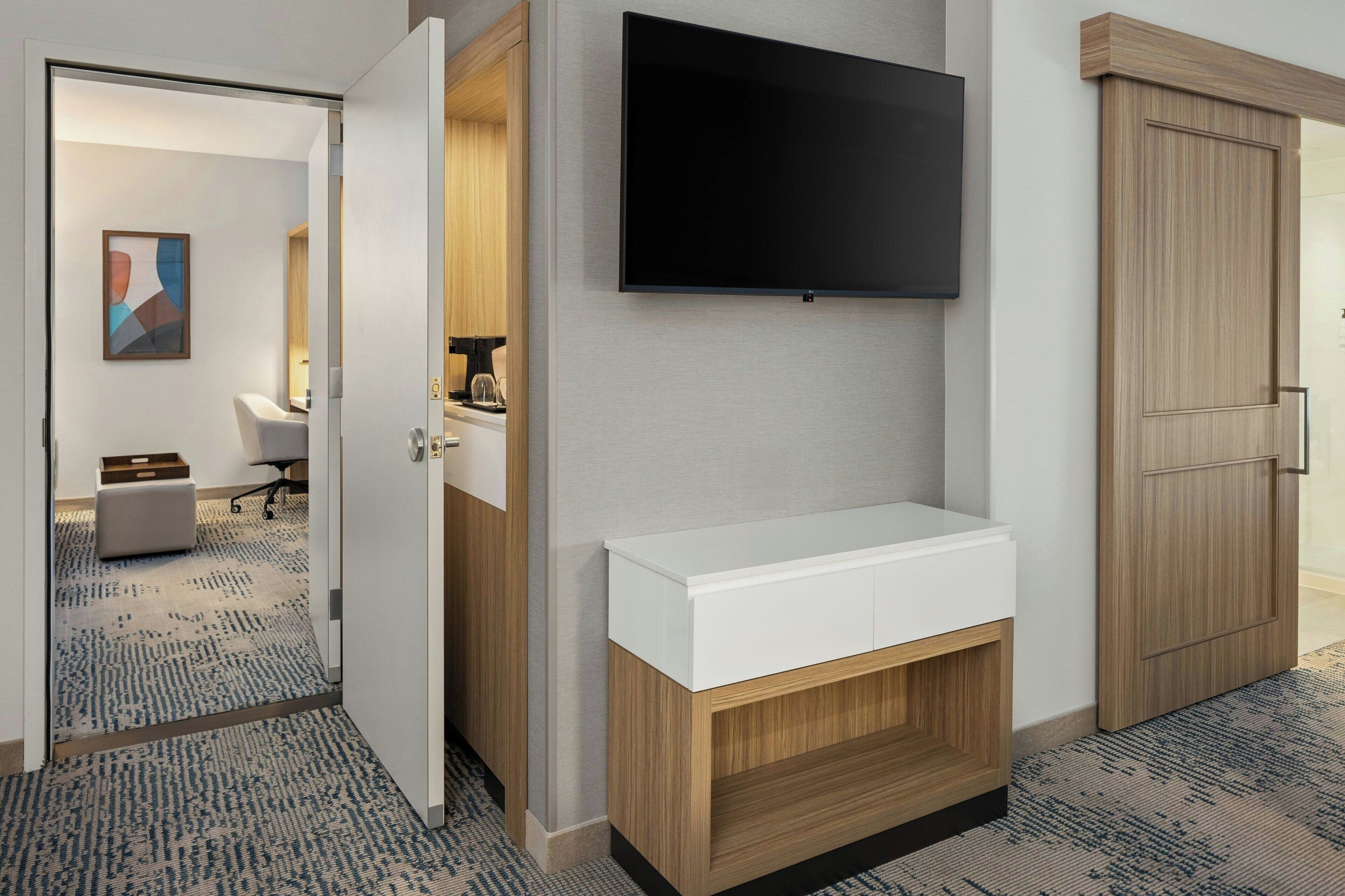 Una habitación de hotel con una televisión de pantalla plana montada en la pared, una mesita de noche blanca y un armario de madera.