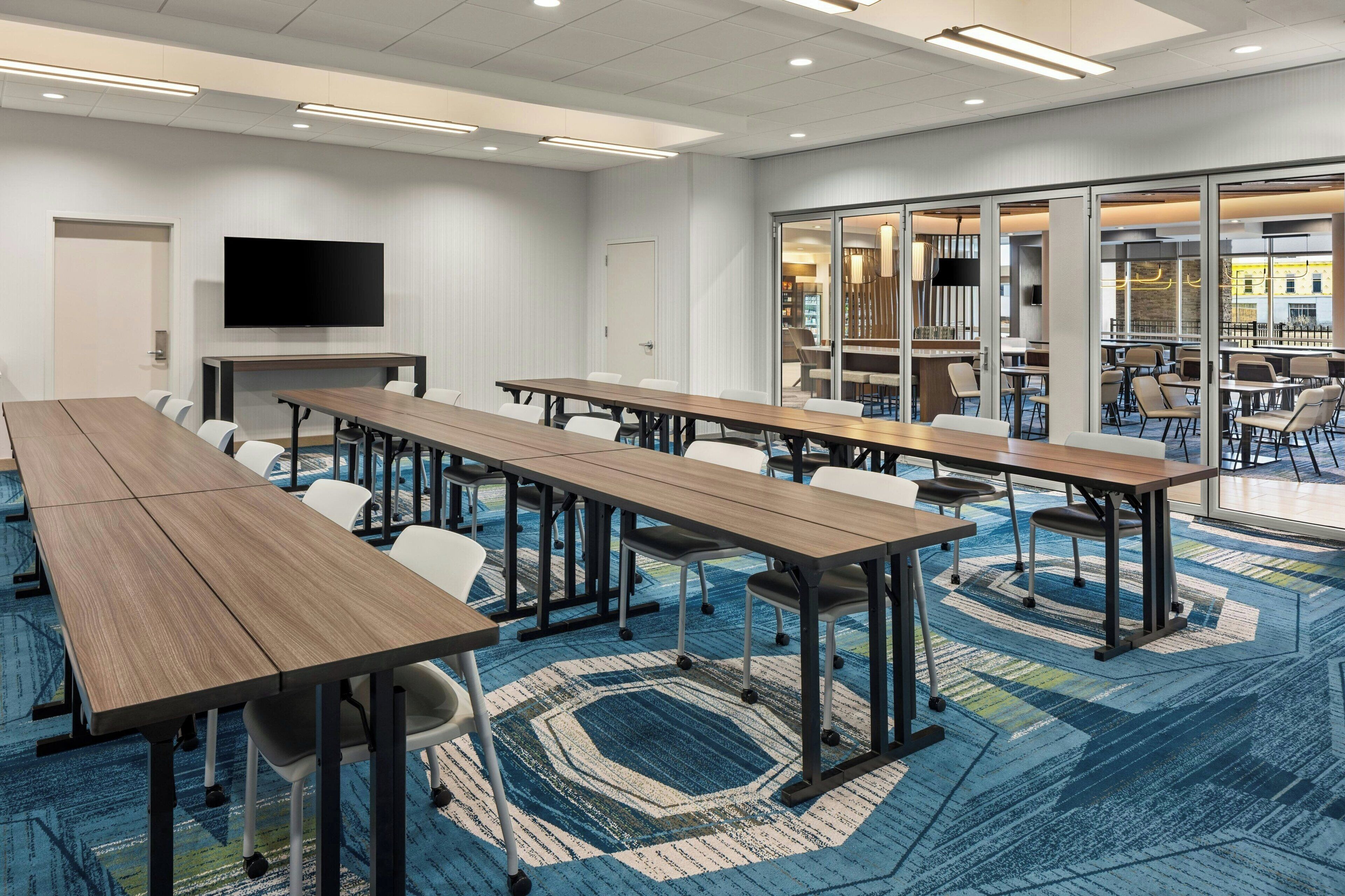 A conference room with long tables and chairs, a flat-screen TV, and a view of a dining area through glass doors.