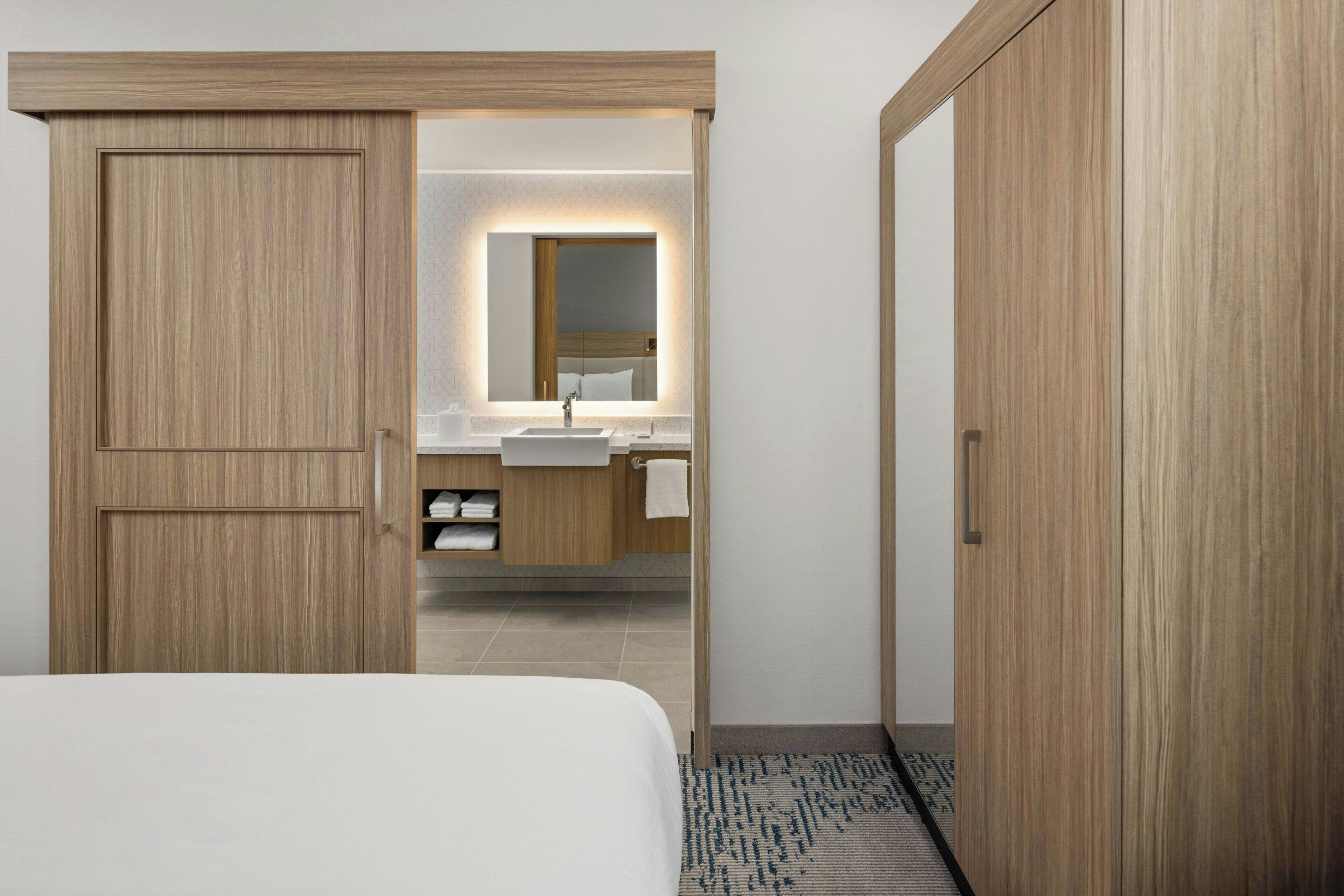 Habitación de hotel con un armario de madera, una cama, un baño con lavamanos y espejo, y una alfombra estampada.