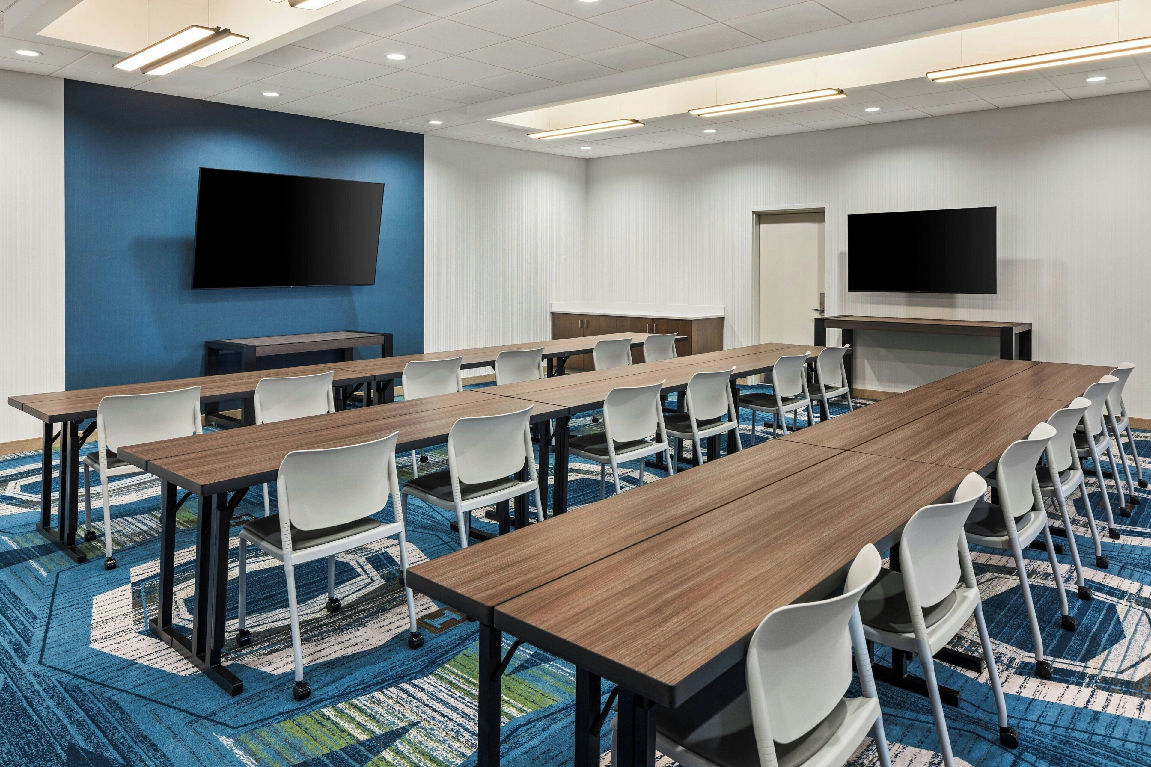 A conference room with long tables, white chairs, a large flat-screen TV, and a patterned carpet.