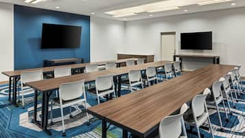 A conference room with long tables, white chairs, a large flat-screen TV, and a patterned carpet.