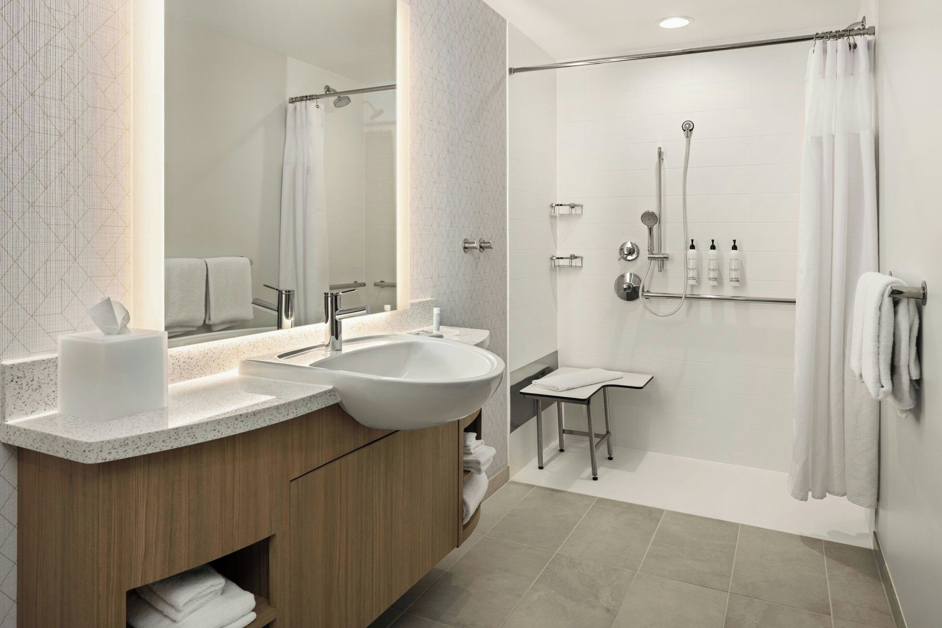 A modern bathroom with a double sink vanity, a large mirror, a shower with a glass door, and a small stool.
