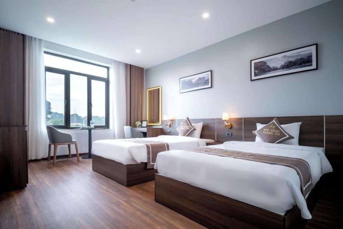 A hotel room with two beds, a desk, and a chair. There are two windows and a view of the city.