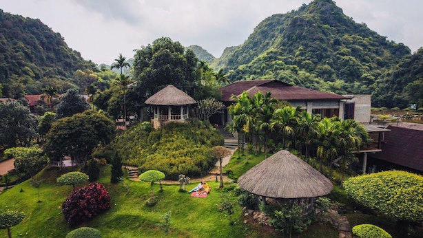 A lush garden with thatched-roof structures, a pathway, and a small pavilion. - Núi Vàng Tràng An (Hoa Lu)