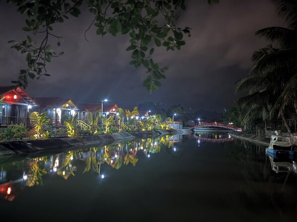 Vista nocturna de una propiedad frente al agua con edificios iluminados, una superficie acuática tranquila y un puente.