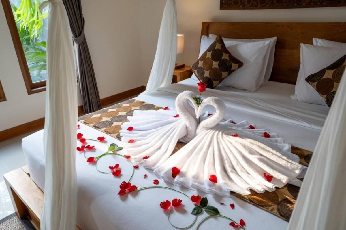 Una cama impecablemente hecha con dos toallas dispuestas en forma de cisnes, un edredón a cuadros y una sola rosa roja.