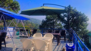 Un espace extérieur aménagé avec des tables et des chaises, un parasol vert et une vue sur les arbres et les collines.
