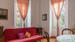 A room with a red sofa, two windows with sheer curtains, a framed picture on the wall, and a dining table with a white tablecloth. - GuestHost - Accommodation of 90 sqm on the first floor of a building with elevator, about 10 steps to access (NOT suitable for disabled), ideal for 4 guests.Located in a strategic position two passes from the Vatican and the Sala Nervi, just steps from S (Roma)