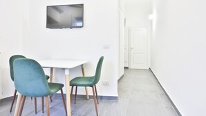 Une pièce avec une table blanche et deux chaises vertes, un téléviseur à écran plat accroché au mur et une porte blanche à l’arrière-plan.