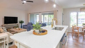 A modern kitchen with a central island, a dining area with wooden chairs, a flat-screen TV, and a ceiling fan. - Lagoon Views, Dog Friendly & Beautifully Renovated - Perfect for a Couple's Beach Getaway! (Kiawah Island)