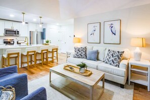 Uma sala de estar moderna com sofá, mesa de centro e cadeiras. Há uma cozinha com micro-ondas, geladeira e banquetas.