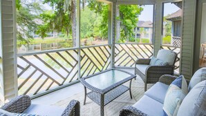 A covered patio with wicker furniture, a glass-top table, and a view of a residential area with trees and houses. - Lagoon Views, Dog Friendly & Beautifully Renovated - Perfect for a Couple's Beach Getaway! (Kiawah Island)