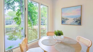 A dining area with a round table, white placemats, wicker chairs, a potted plant, and a painting of a beach scene. - Lagoon Views, Dog Friendly & Beautifully Renovated - Perfect for a Couple's Beach Getaway! (Kiawah Island)