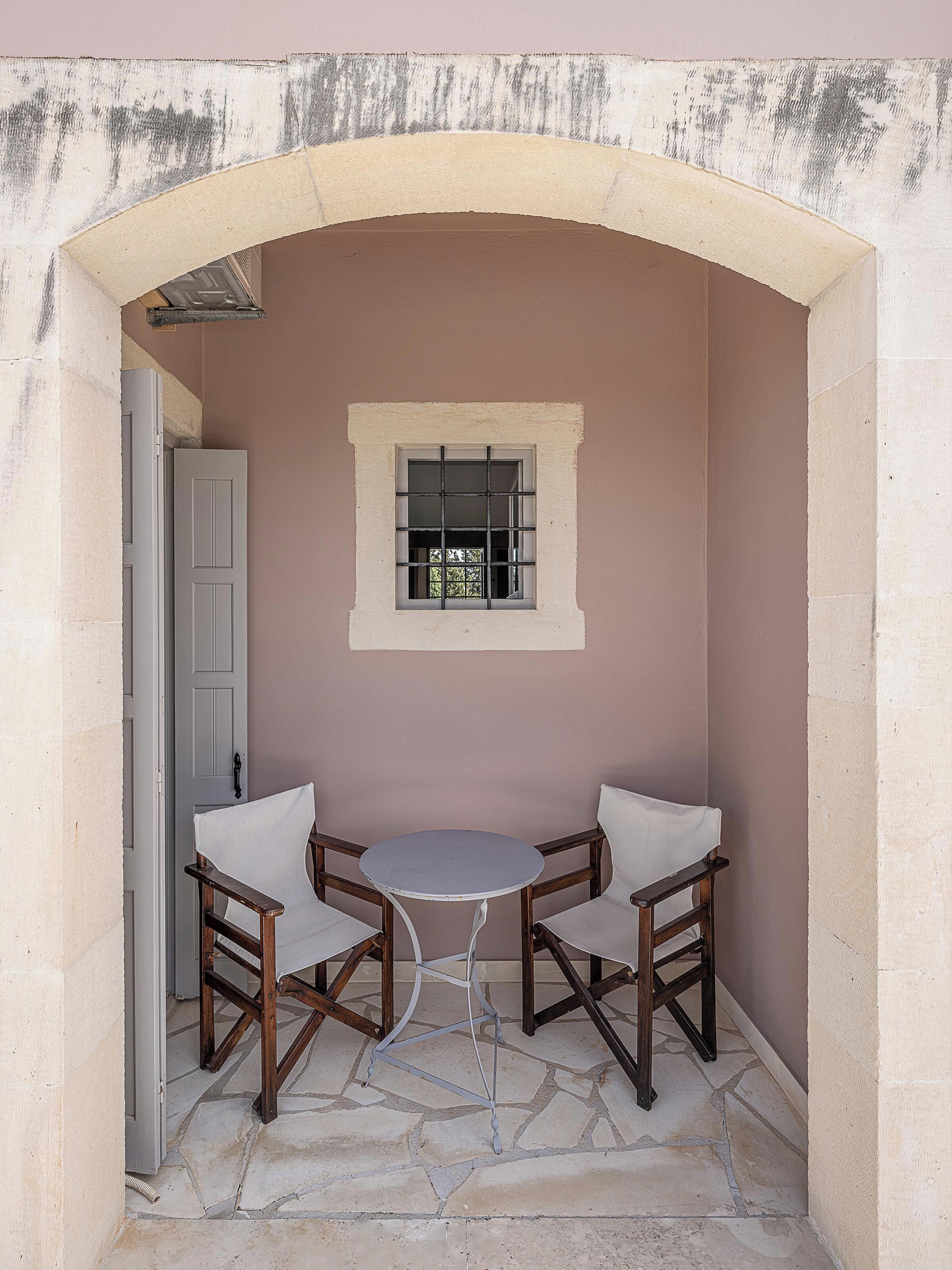 Un petit espace extérieur aménagé avec deux chaises et une table, délimité par une arche en pierre et une fenêtre à barreaux.