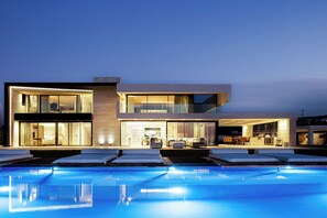 A modern two-story house with a swimming pool in front, featuring a dining area and a kitchen visible through the glass doors.