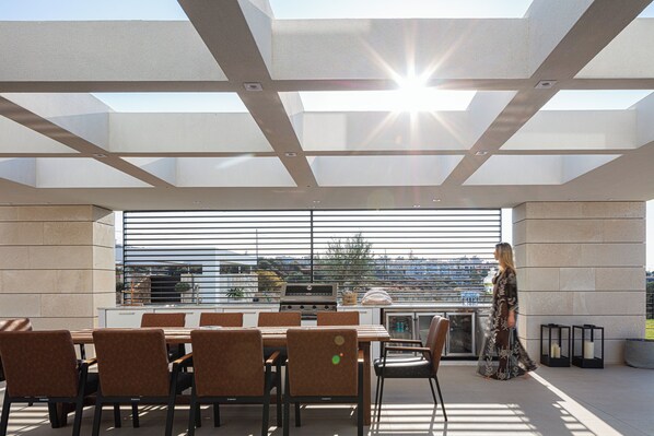 A modern outdoor dining area with a large table and chairs, a grill, and a view of the cityscape.