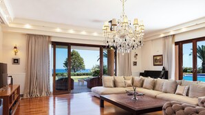 Ein geräumiges Wohnzimmer mit einer großen Couch, einem Couchtisch und einer Chandelier. Ein Klavier steht dort, und große Fenster bieten Blick ins Freie.