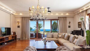 Ein geräumiges Wohnzimmer mit einer großen Couch, einem Couchtisch und einer Chandelieber. Ein Fernseher steht auf einem Holzboden, und das Zimmer verfügt über große Fenster mit Meerblick.