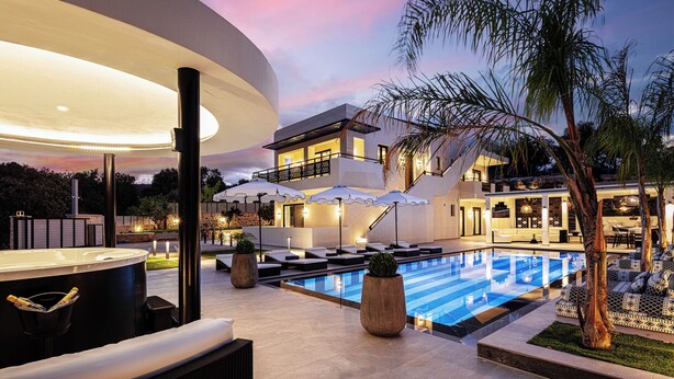 A modern outdoor pool area with lounge chairs and umbrellas, surrounded by a well-maintained garden and a large building with a balcony overlooking the pool.