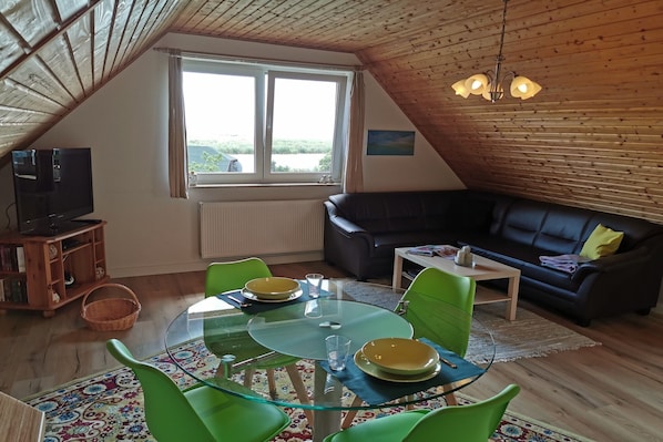 Ein gemütliches Wohnzimmer mit einem Glastisch, grünen Sesseln, einem schwarzen Sofa und einem Fernseher auf einer Holzkommode.