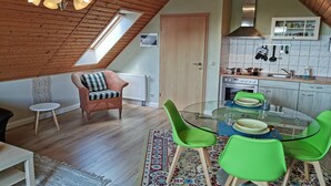 A cozy loft-style room with a sloped ceiling, a dining area with a glass table and green chairs, a kitchenette with white cabinets, and a wooden ceiling. - Charming Attic Retreat, Usedom (Usedom)