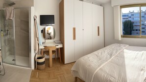 Un dormitorio con cama, escritorio, silla, televisión y ventana con vista a edificios.