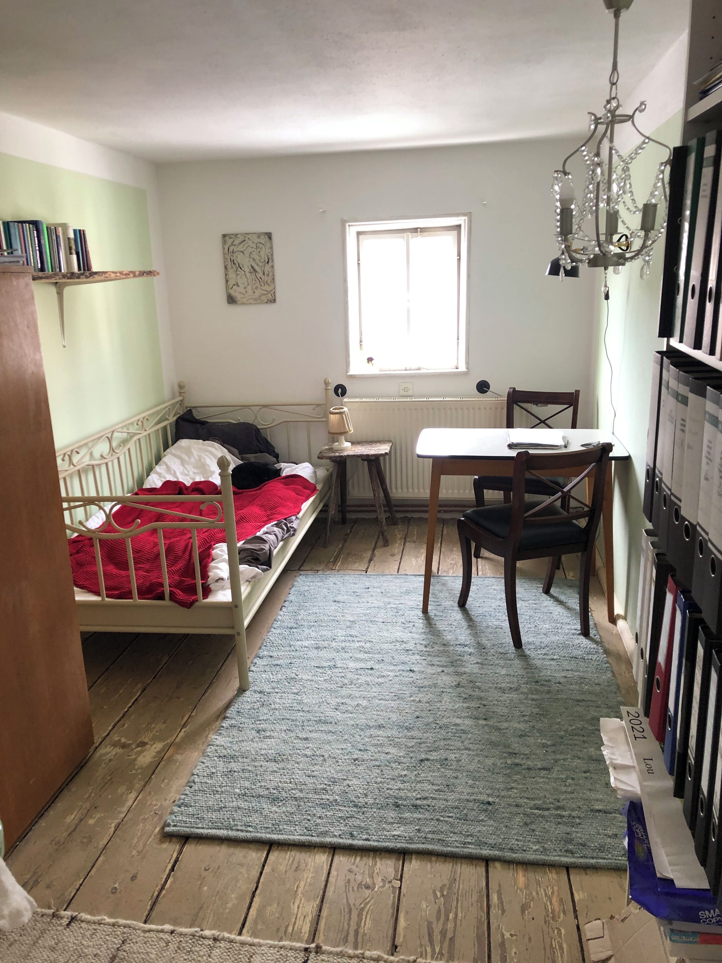 A small, cozy room with a single bed, a small table, a chair, a radiator, a window, and a bookshelf.