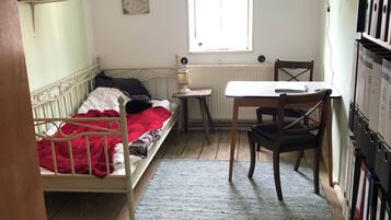 A small, cozy room with a single bed, a small table, a chair, a radiator, a window, and a bookshelf.