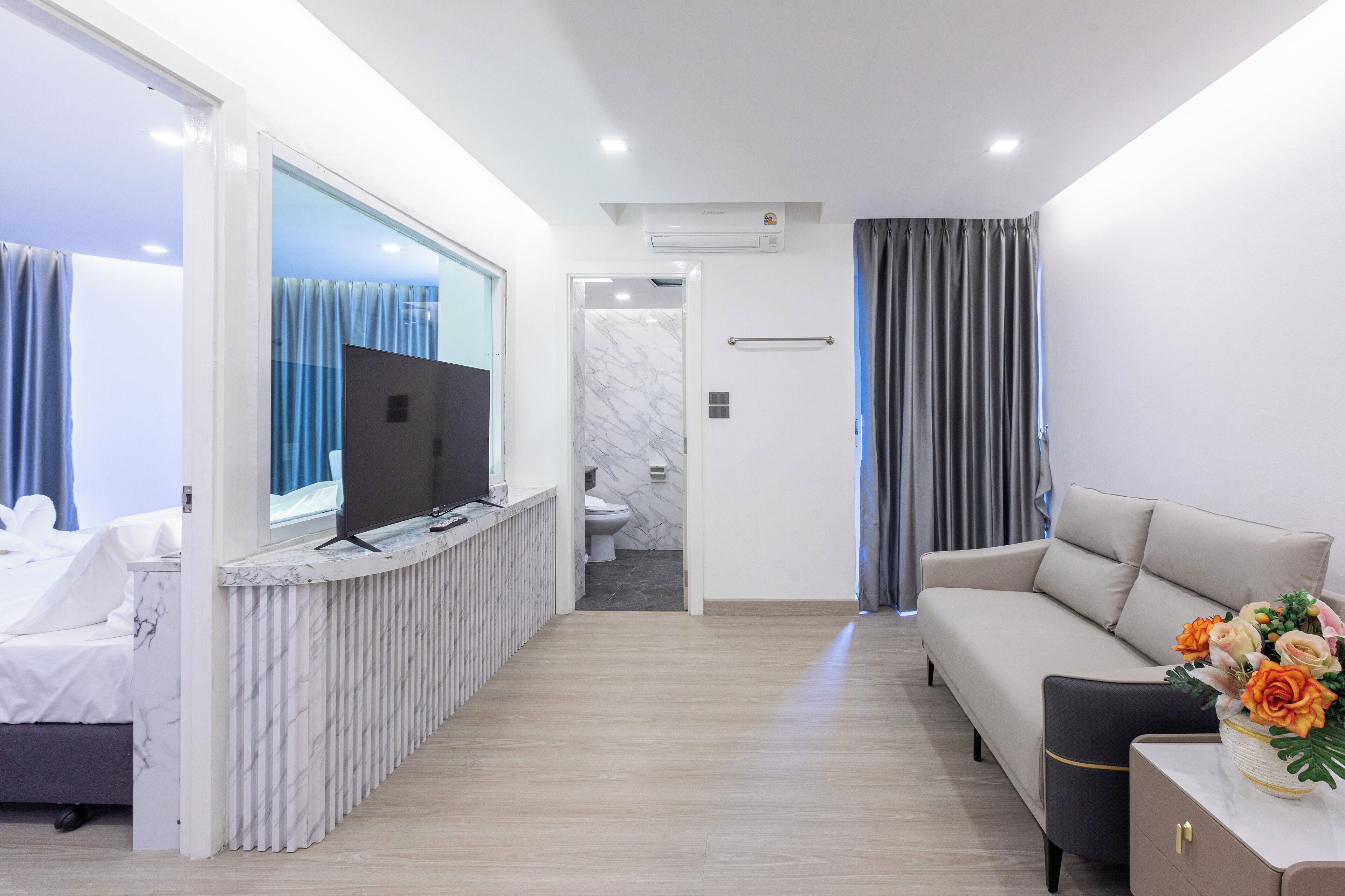 A modern hotel room with a bed, a TV, a sofa, and a bathroom visible through an open door.