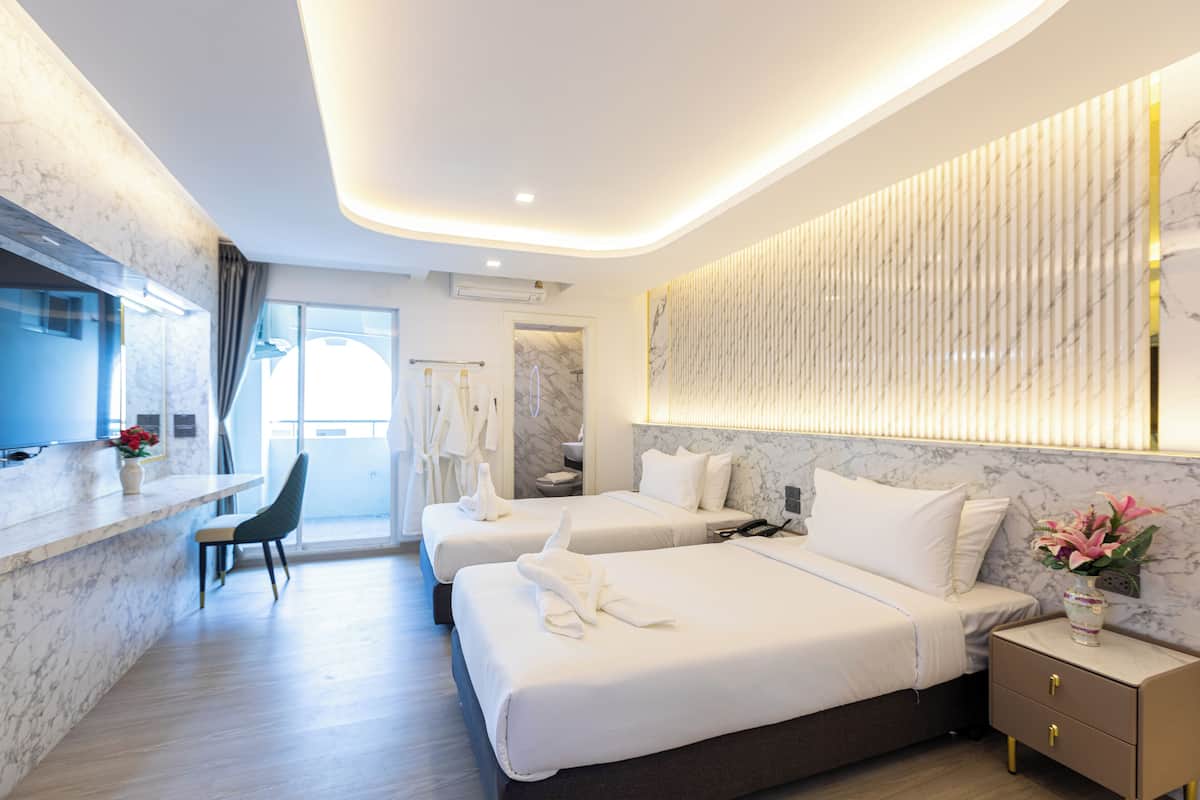 Una habitación de hotel moderna con dos camas, una zona de estar con una silla, un escritorio pequeño y un baño visible a través de una puerta abierta.