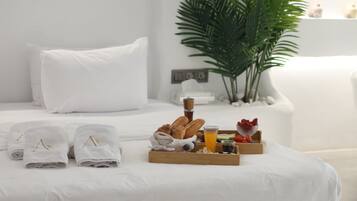 Ein ordentlich bezogenes Bett mit weißen Leinentüchern, ein Frühstücksbrett mit Croissants, Obst und Saft, und eine Topfpflanze im Hintergrund.