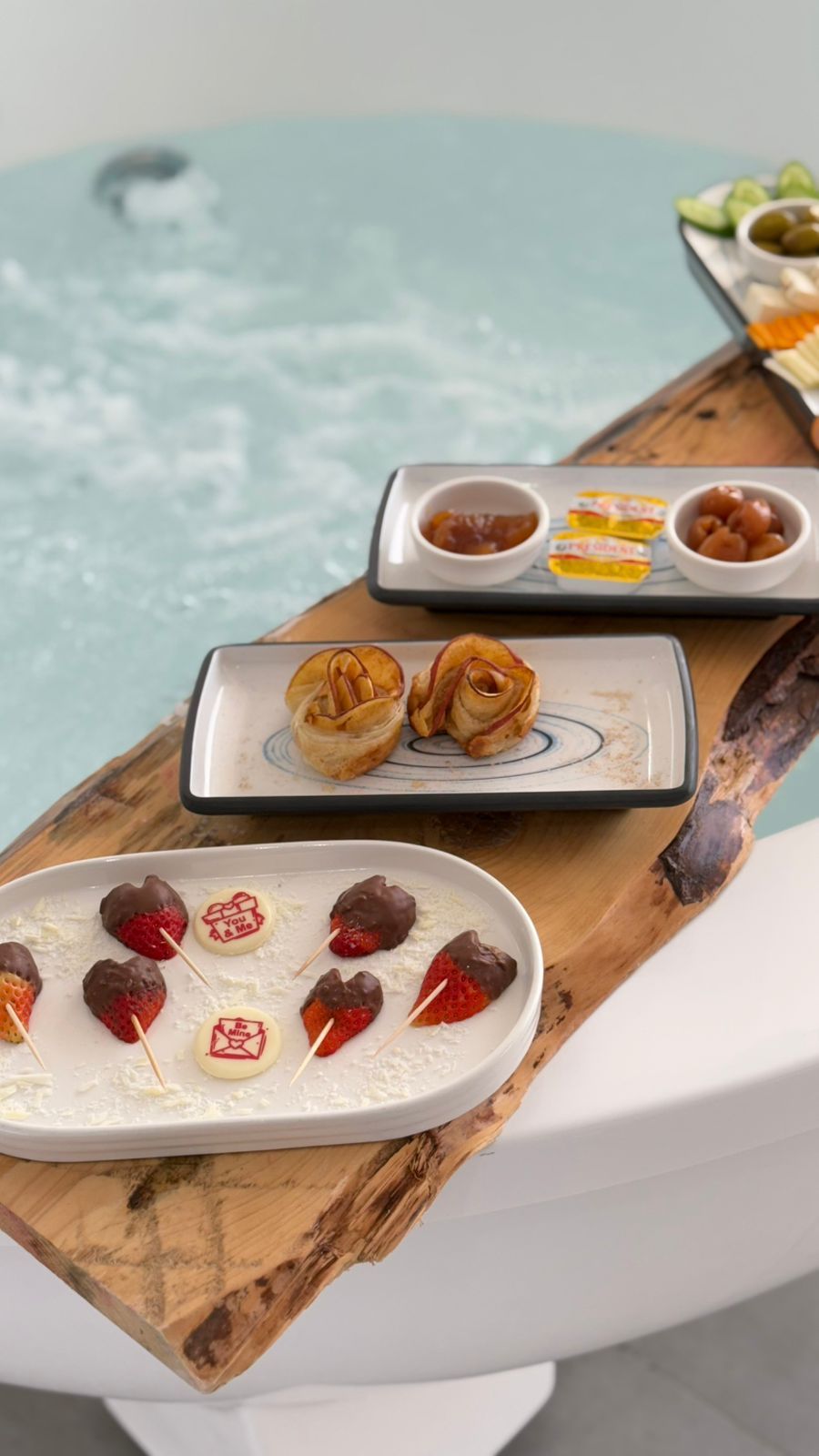 A tray of food items including pastries, chocolate-covered strawberries, and a bowl of olives, placed on a wooden board near a hot tub.