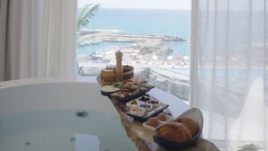 Una habitación con una bañera grande, una bandeja de desayuno con pasteles y bebidas, y vistas al mar y barcos.