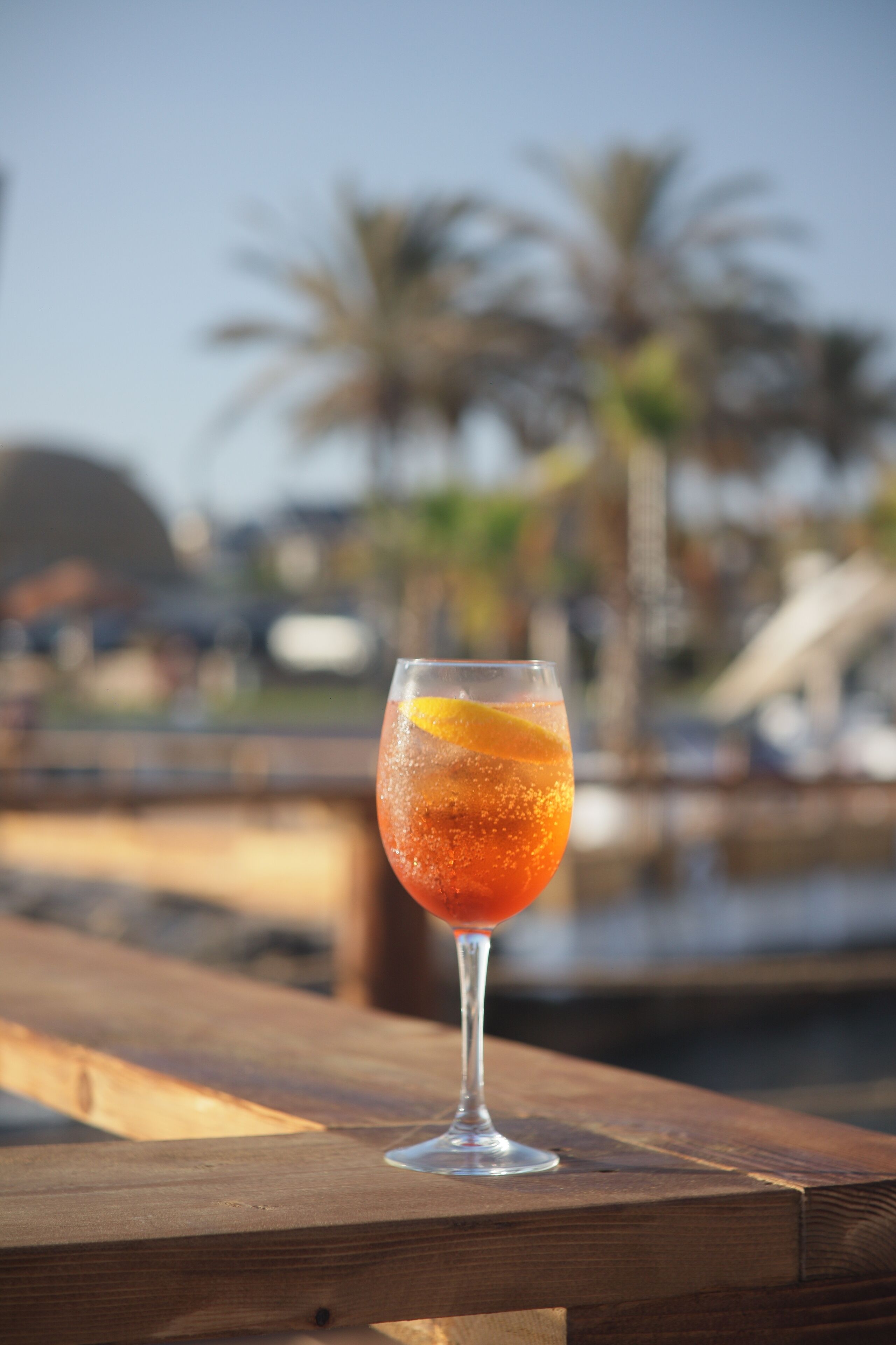A glass of red beverage with an orange slice on a wooden table with a marina and palm trees in the background.